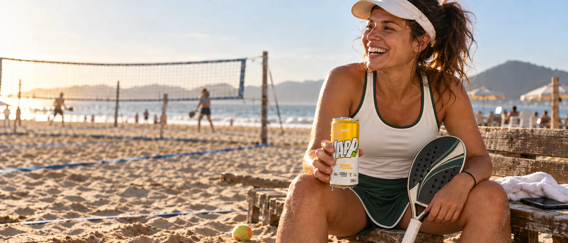 Mulher sorrindo com uma lata de YAPP! Manga com Maracujá e uma raquete de beach tennis na praia